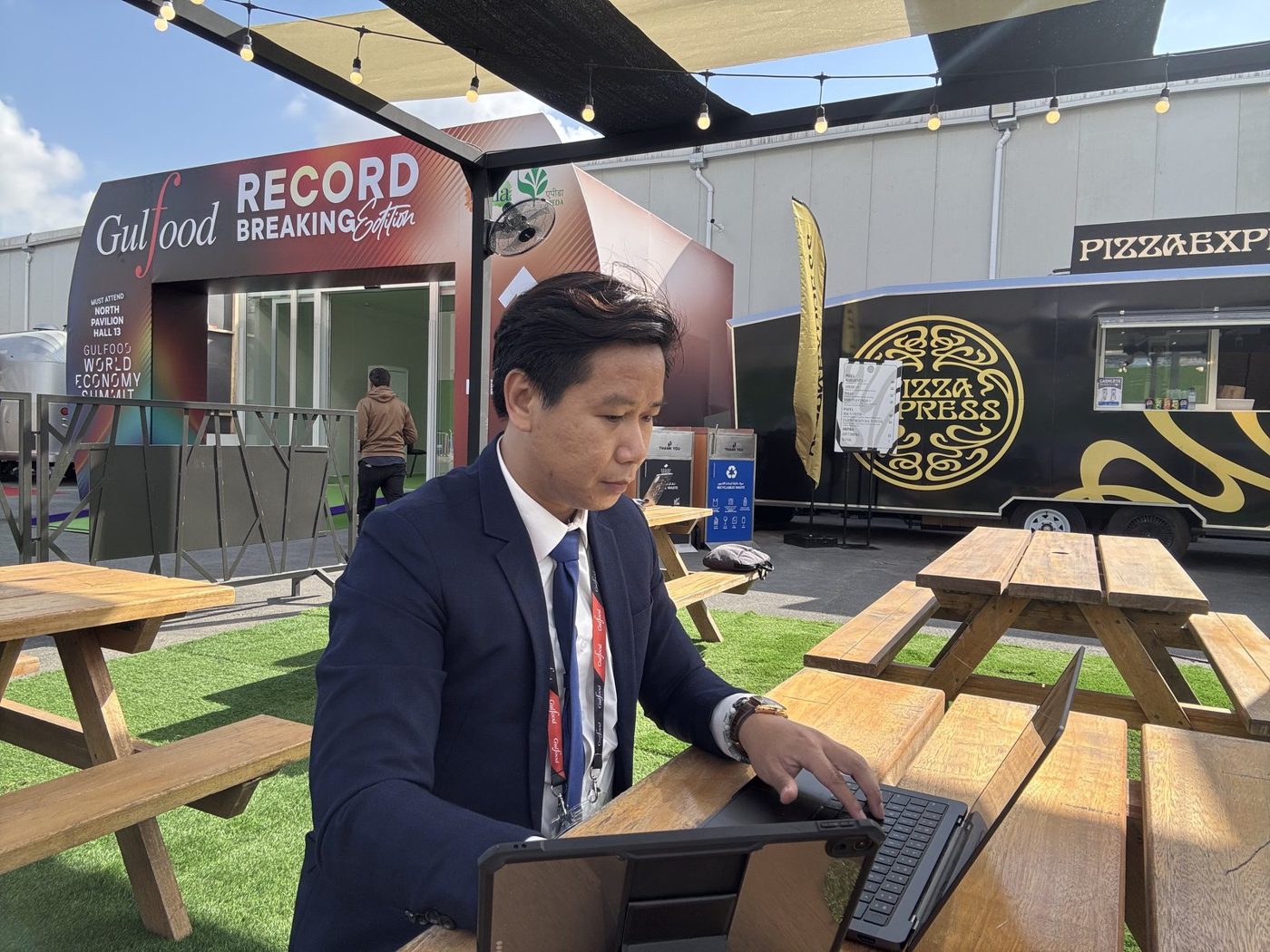 Maximillien Quân Phạm làm việc bằng laptop tại bàn ngoài sân Gulfood 2026 vì thiếu chỗ ngồi tiếp khách